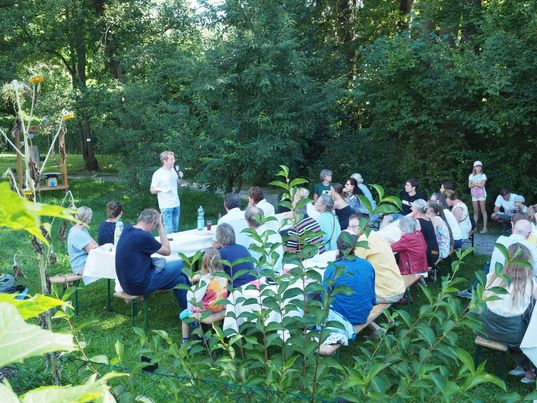 Vortragender steht vor einer Gruppe von Personen, die in einem Garten auf Bänken sitzen und ihm zuhören 