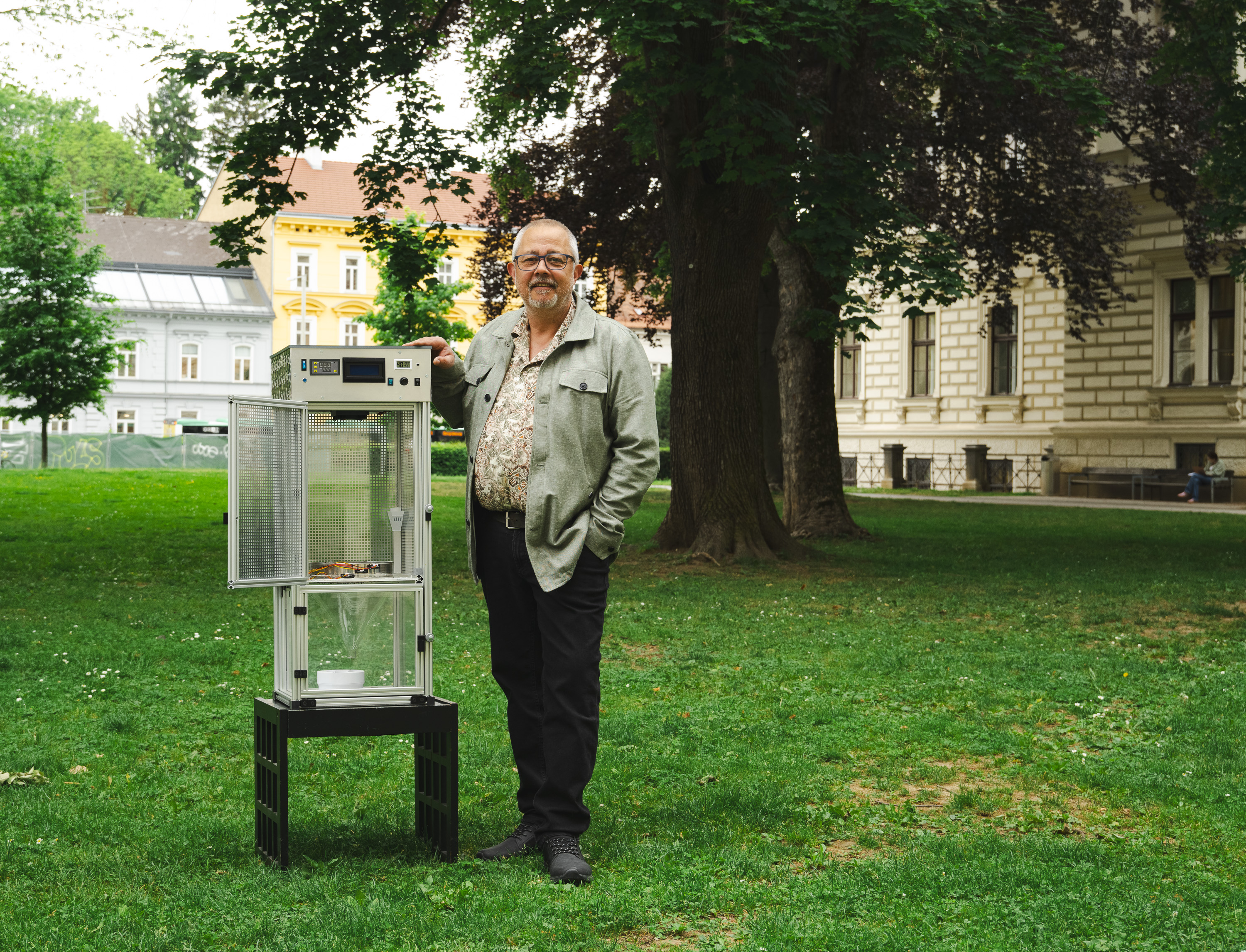 Ein Mann mittleren Alters steht neben einer offenen Glasvitrine auf der Wiese eines Parks auf dem Campus der Universität Graz. Die Vitrine hat er unter den Arm geklemmt. Er lächelt leicht in die Kamera und posiert für ein Foto