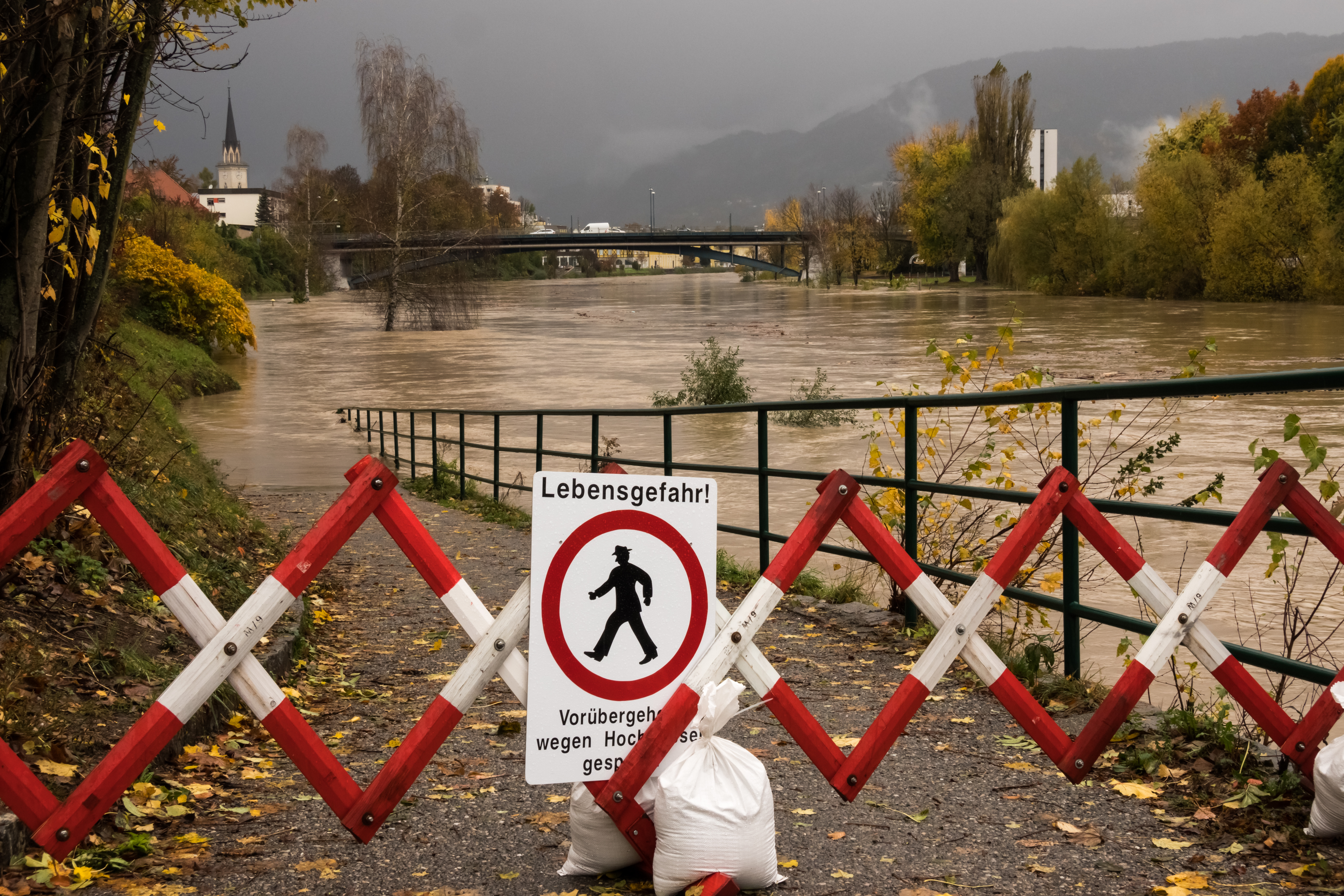 Ein Schild mit der Aufschrift „Lebensgefahr“ wird während des Herbsthochwassers vor einem rot-weiß gestreiften Eisenzaun am überfluteten Fluss Drau in Villach, Österreich, auf den Boden gelegt. 