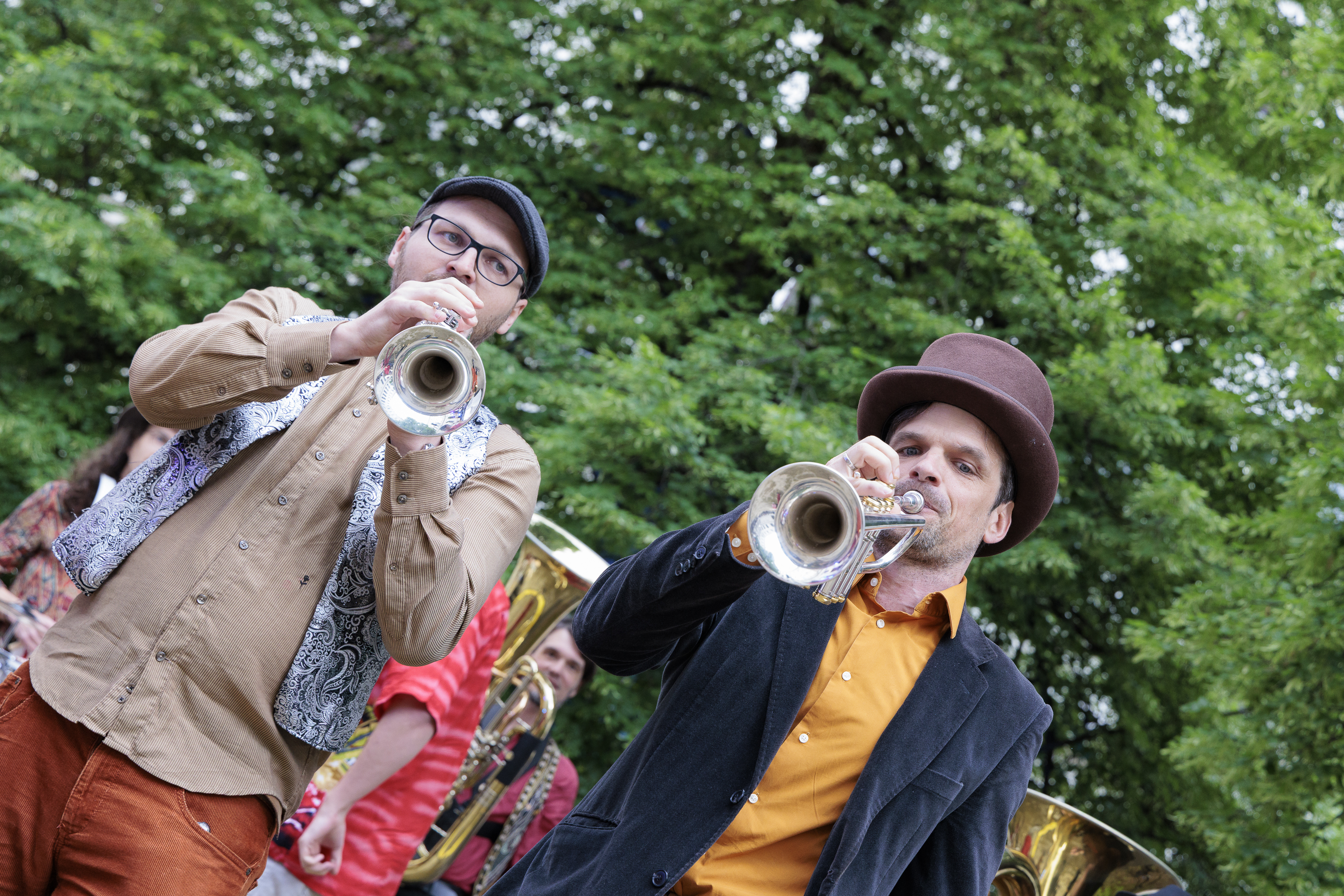 Zwei Männer im Park, einer spielt Posaune, der andere Tuba, beide tragen braune Fedora-Hüte, sind leger gekleidet und stehen auf einer Party im Freien vor dem Hintergrund grüner Bäume. ©Uni Graz/Harry Schiffer