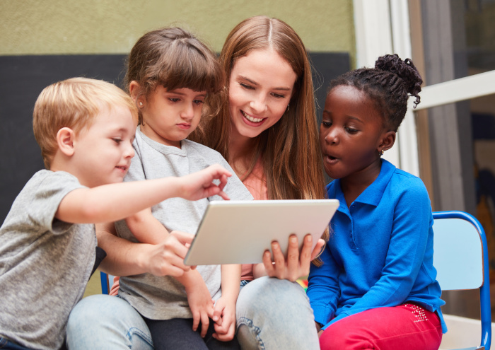 Decoration: A smiling young woman sits with three children, who are looking attentively at a tablet she is holding in her hands. ©Robert Kneschke/Adobe Stock