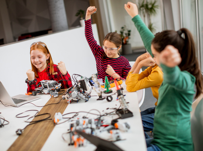 Dekoration: Kinder heben begeistert die Arme hoch, während sie vor einem Laptop sitzen und mit Robotik-Bausätzen auf einem Tisch arbeiten. ©BGStock72/Adobe Stock