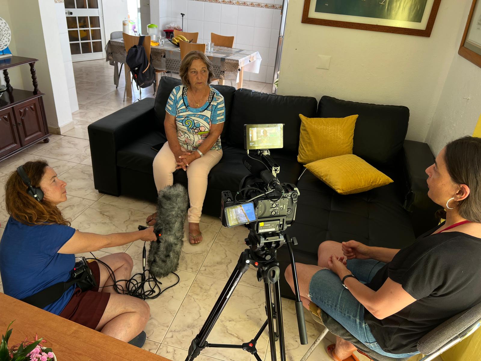 Three women are filming an interview in a living room with a video camera, microphones, and sound equipment. One woman is seated on a black couch being interviewed. ©Rodrigo Lacerda