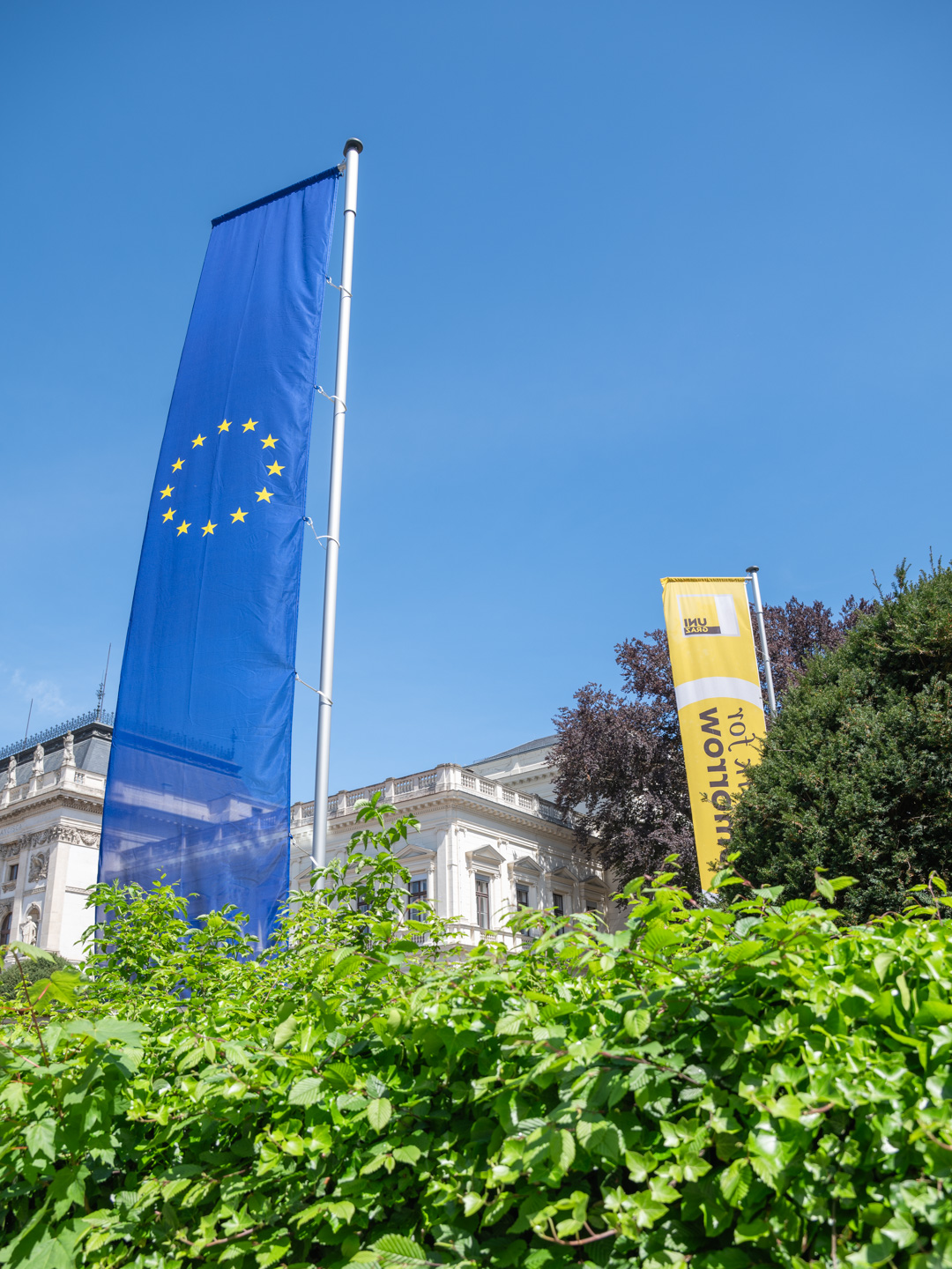 Eine europäische Flagge und ein Werbebanner an einem öffentlichen Gebäude vor einer Hecke. Foto: Uni Graz/Angele ©Uni Graz/Angele