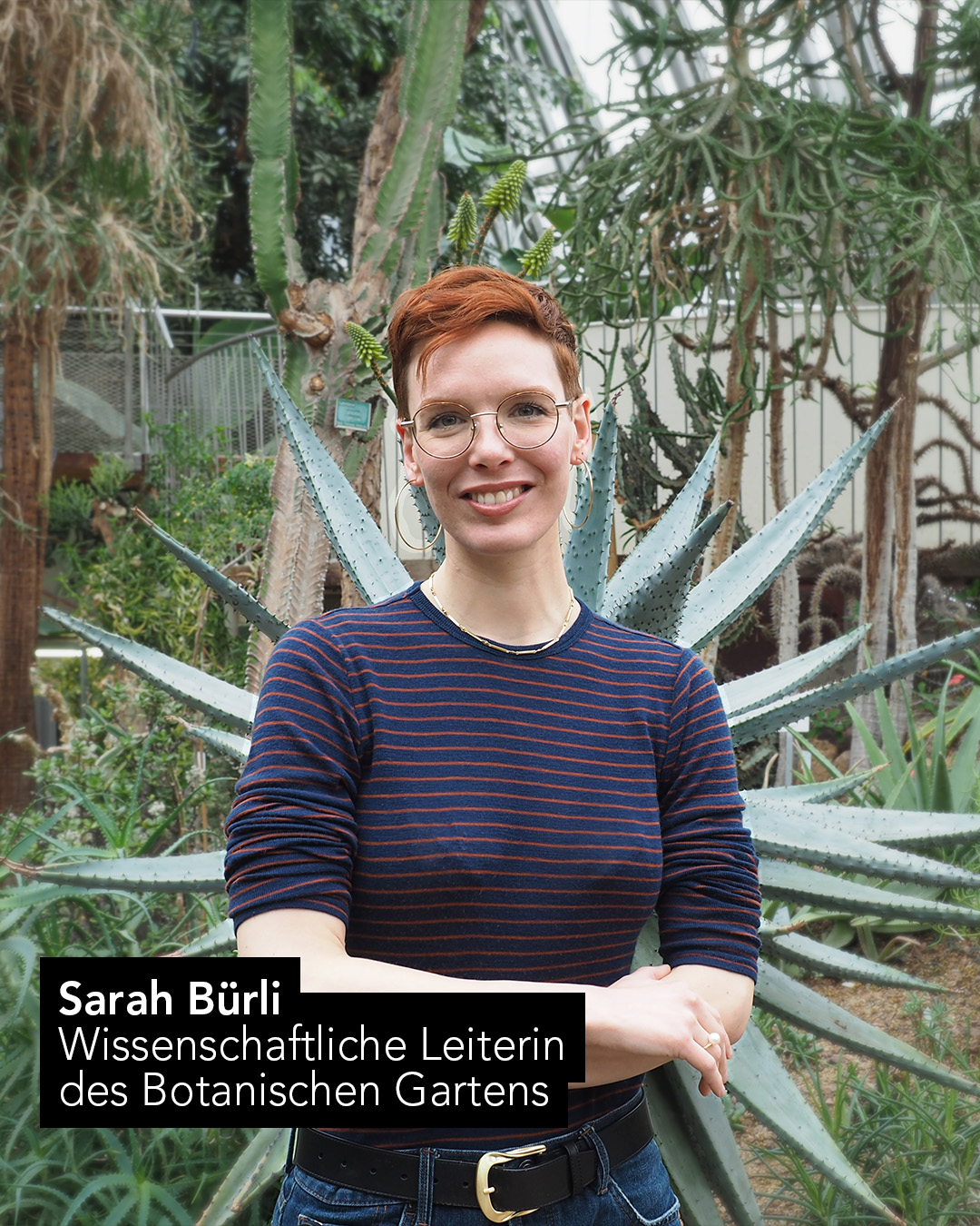 Eine Person mit kurzen Haaren steht mit verschränkten Armen vor großen Sukkulenten und Kakteen in einem botanischen Garten; im Vordergrund ist ein Namensschild mit der Aufschrift „Sarah Bürli, Wissenschaftliche Leiterin des Botanischen Gartens“ zu sehen. 