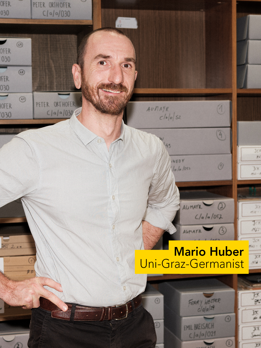 Eine Person mit weißem Hemd steht vor einem Regal mit beschrifteten Archivboxen. Die Boxen sind ordentlich gestapelt, und die Umgebung deutet auf ein Archiv oder eine Bibliothek hin. Ein gelbes Textfeld zeigt den Namen "Mario Huber" und die Bezeichnung "Uni-Graz-Germanist". 