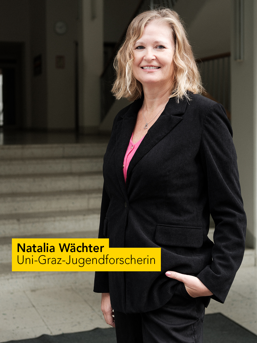 Eine Person steht in einem Innenbereich vor einer Treppe. Sie trägt einen schwarzen Blazer und ein pinkfarbenes Oberteil. Im Hintergrund sind eine Treppe, Wände und eine Uhr zu sehen. Ein gelber Textblock enthält die Aufschrift „Natalia Wächter, Uni-Graz-Jugendforscherin“. 