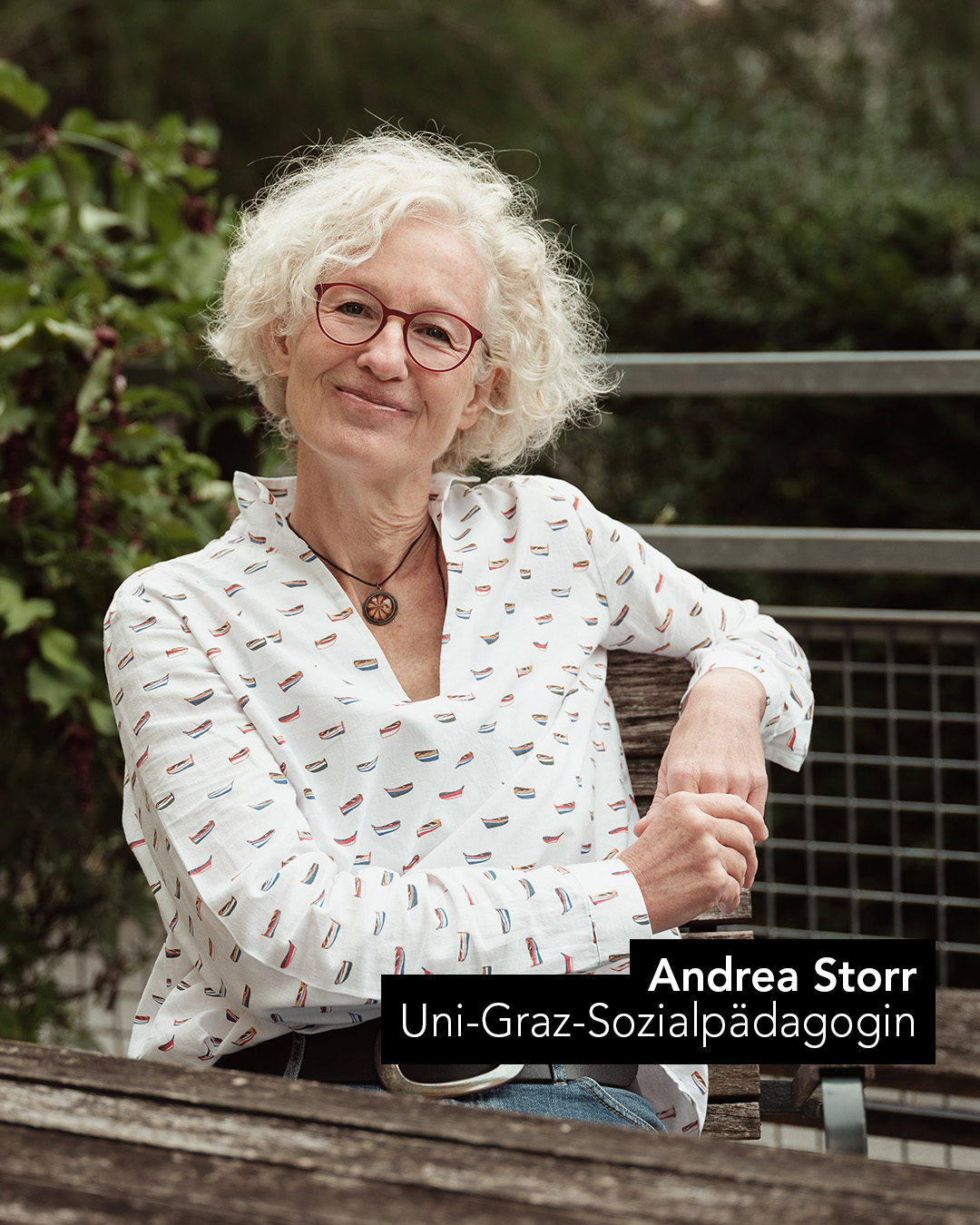 Ein Frau mit weißen Haare und einem gemusterten weißen Oberteil sitzt an einem Holztisch mit weißem Hintergrund 