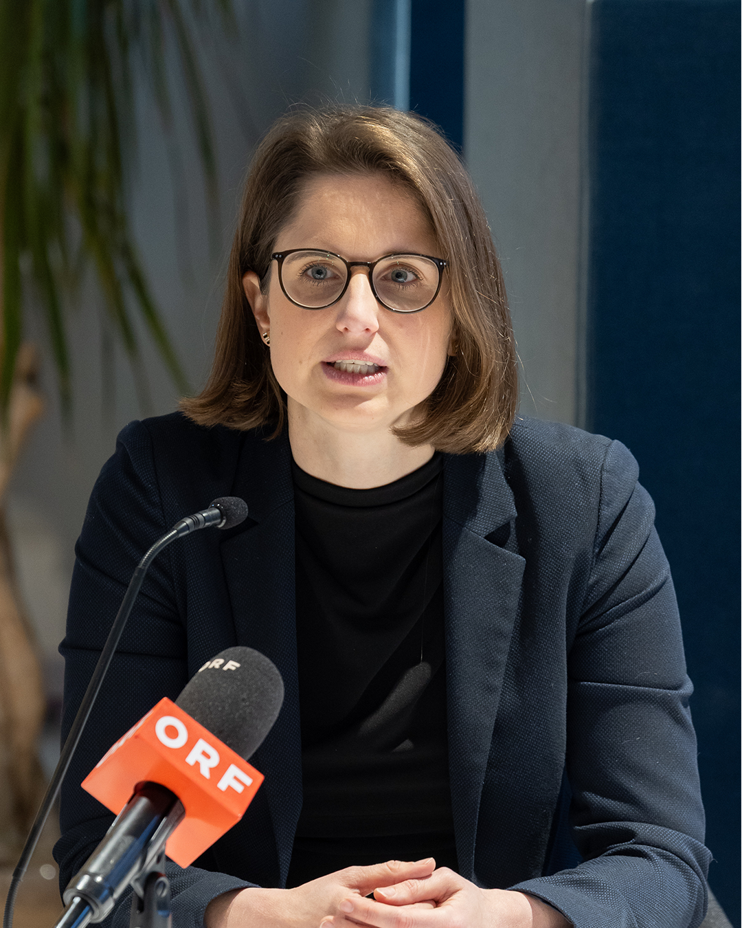 Eine Person in einem dunklen Blazer sitzt vor einem Mikrofon mit ORF-Logo. Im Hintergrund sind unscharfe Details wie eine Pflanze und eine blaue Wand zu erkennen. Die Szene wirkt wie eine professionelle Gesprächs- oder Interview-Situation. 