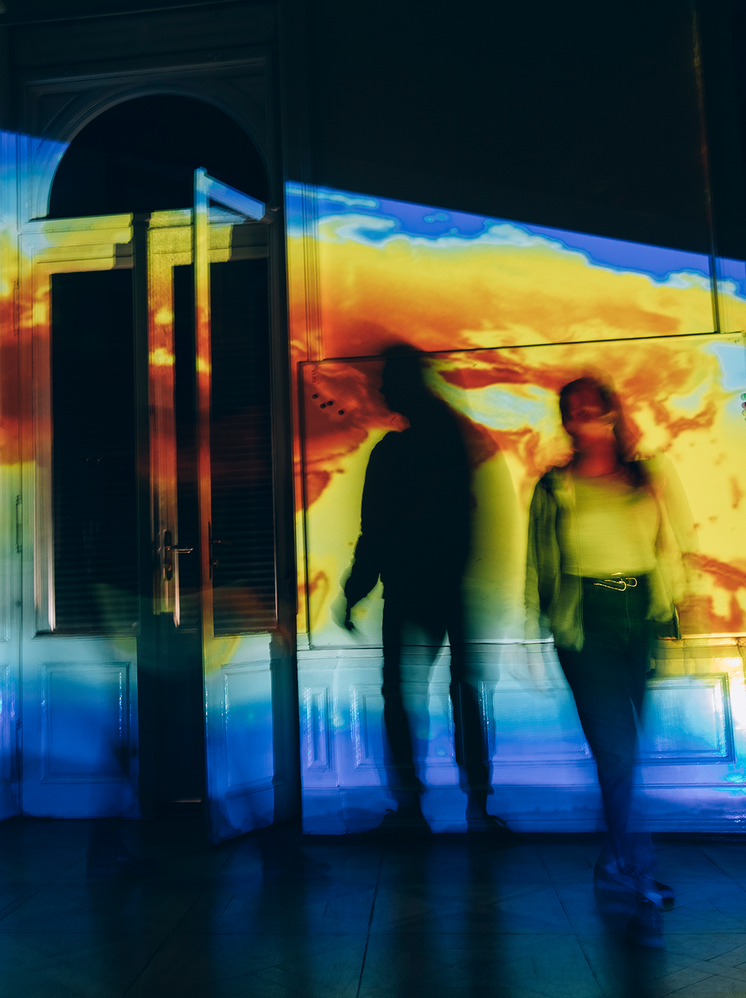 Zwei unscharfe, schemenhafte Personen stehen vor einer Wand, auf der bunte Lichtprojektionen in Gelb, Blau und Orange zu sehen sind. Im Hintergrund ist eine Tür erkennbar. 