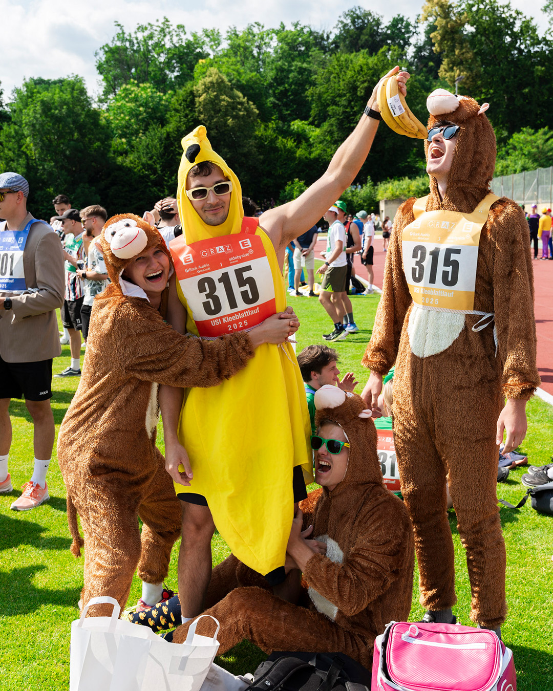 Barrierefreier Alternativtext: Mehrere Personen stehen und sitzen auf einer Wiese bei einer Sportveranstaltung. Drei von ihnen tragen braune Tierkostüme mit weißen Bäuchen, die an Affen erinnern. Eine Person trägt ein gelbes Bananenkostüm und hält einen Bund Bananen in die Luft. Alle tragen Startnummern mit der Nummer 315 und dem Schriftzug „USI Kleeblattlauf 2025“. Im Hintergrund sind weitere Teilnehmende und Zuschauer auf einer Laufbahn zu sehen, umgeben von grünen Bäumen. Vor den Kostümierten stehen Taschen und Rucksäcke auf dem Gras. 