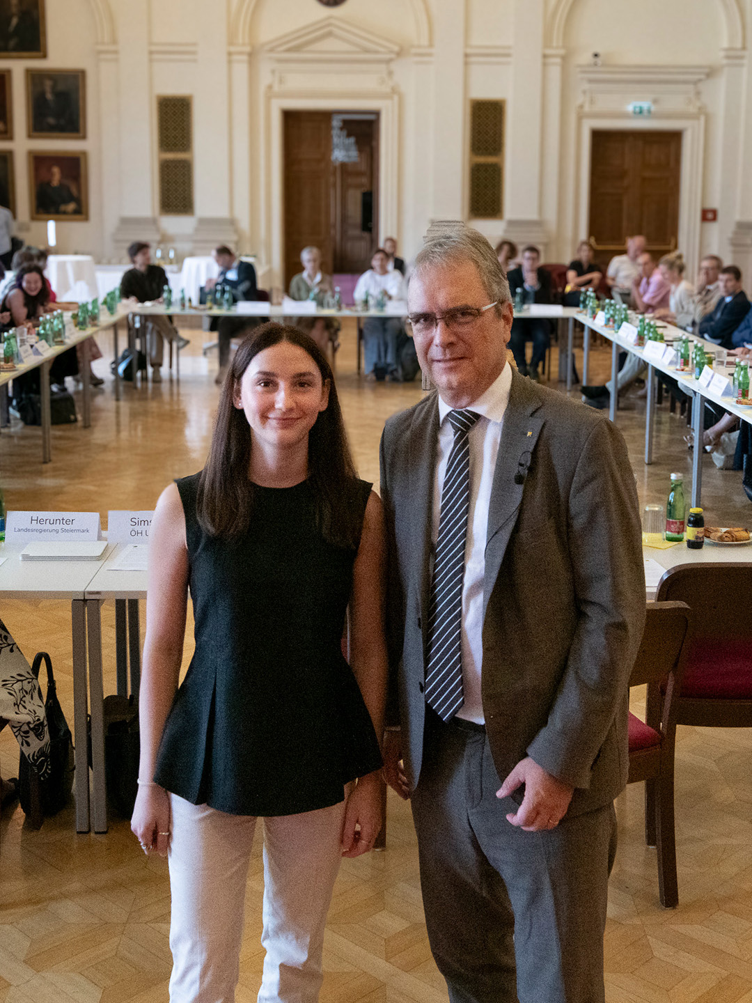 Eine Frau mit schwarzem Top und ein Mann in grauem Anzug vor einer im Viereck aufgestellten Tisch Reihe in einem großen Raum ©Uni Graz/Tzivanopoulos