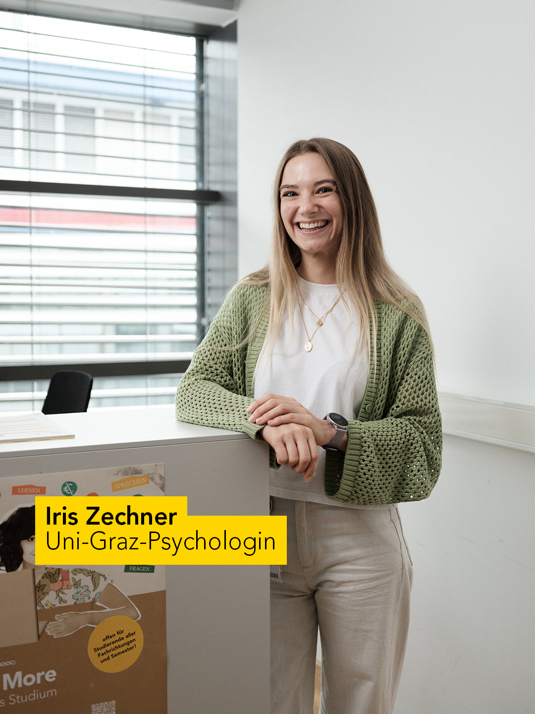 Eine Person steht in einem modernen Raum neben einer weißen Box, die mit bunten Grafiken und Texten dekoriert ist. Die Person trägt eine grüne Strickjacke, ein weißes T-Shirt und helle Hosen. Im Hintergrund sind große Fenster mit Jalousien sichtbar, die viel Tageslicht hereinlassen. Die Szene wirkt freundlich und professionell. Auf der Box ist ein gelber Textblock mit der Aufschrift „Iris Zechner, Uni-Graz-Psychologin“ angebracht. 