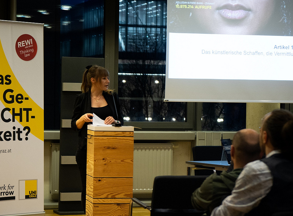 © Uni Graz/Nedovic Antonia Bruneder spricht am Podium der Veranstaltung. Im Hintergrund der Beamer mit einer Darstellung zum Thema © Uni Graz/Nedovic
