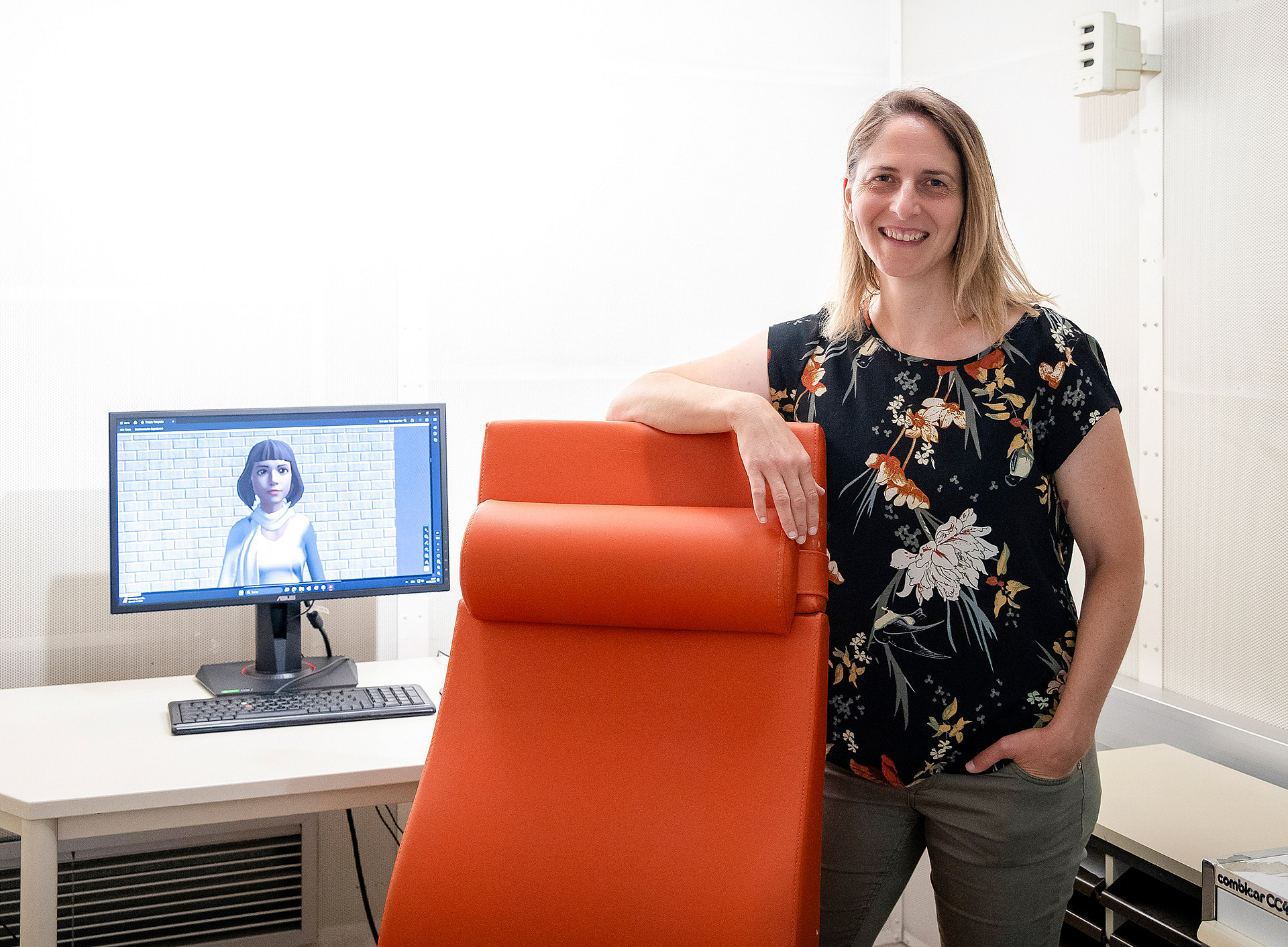 Silvia Kober steht im Psychologie-Labor an einen roten Sessel gelehnt, links im Bild ein Monitor, auf dem ein weiblicher Avatar zu sehen ist ©Uni Graz/Kaiser