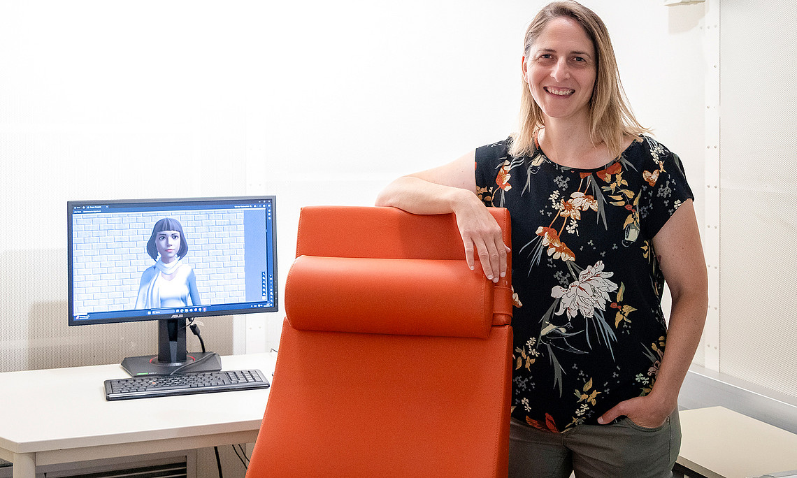Silvia Kober steht im Psychologie-Labor an einen roten Sessel gelehnt, links im Bild ein Monitor, auf dem ein weiblicher Avatar zu sehen ist