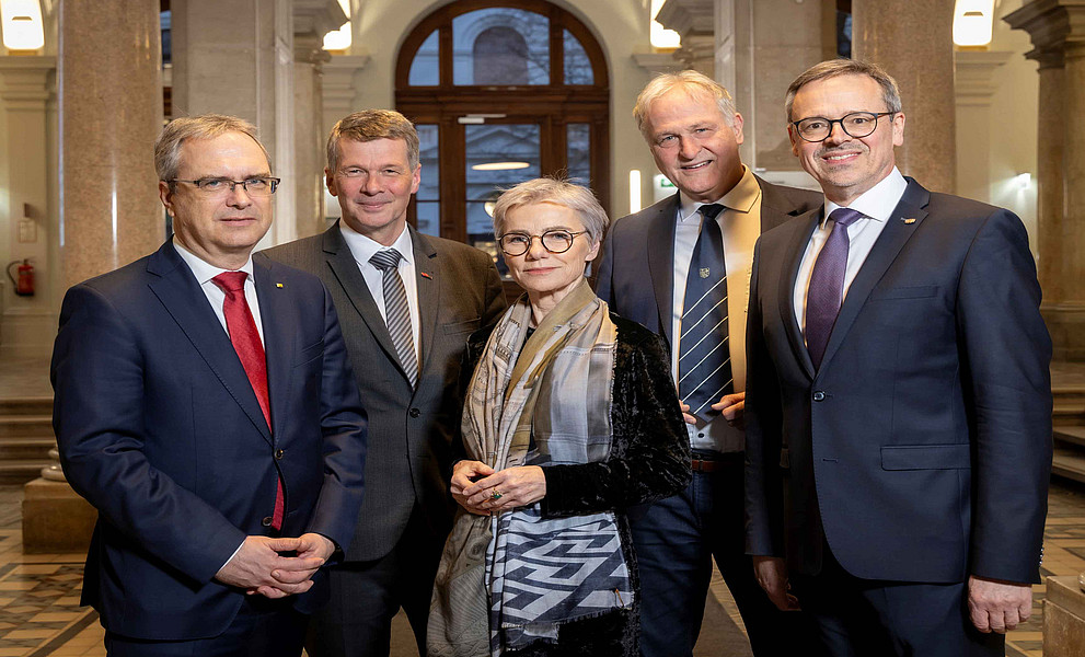 Fünf Erwachsene in formeller Kleidung stehen in einer historischen Eingangshalle mit Säulen und Mosaikboden, eine Person trägt einen Schal.