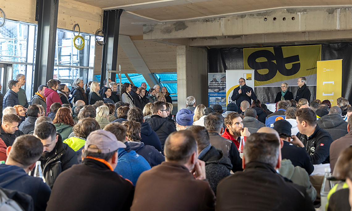 ein Mann spricht im Rohbau eines Dachgeschosses in ein Mikrofon, eine große Menge an Menschen sitzt im Publikum und hört zu