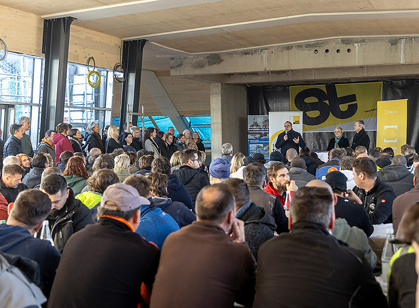 ein Mann spricht im Rohbau eines Dachgeschosses in ein Mikrofon, eine große Menge an Menschen sitzt im Publikum und hört zu ©© Helmut Lunghammer