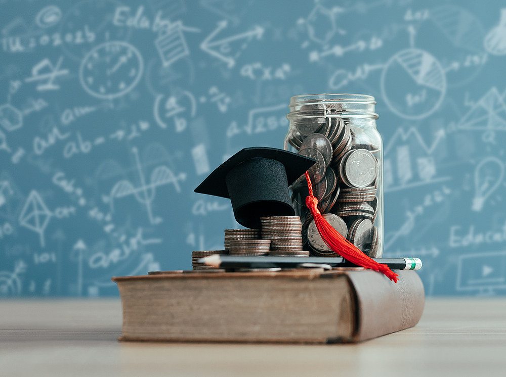 a jar filled with coins on a book together with a small college hat ©By narawit; stock.adobe.com