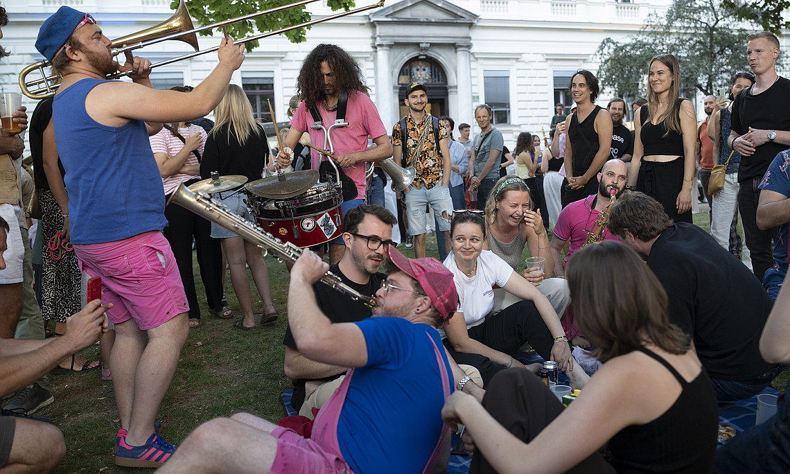 A crowd on the lawn in front of the university building; several people are playing music on a trombone, saxophone and drum, whilst others sit in a circle and listen.