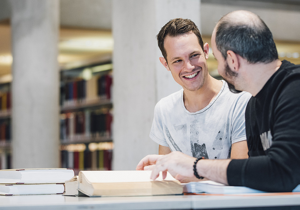 Zwei Studierende sitzen gemeinsam mit Büchern an einem Tisch in der Universitätsbibliothek und lachen und lernen zusammen.