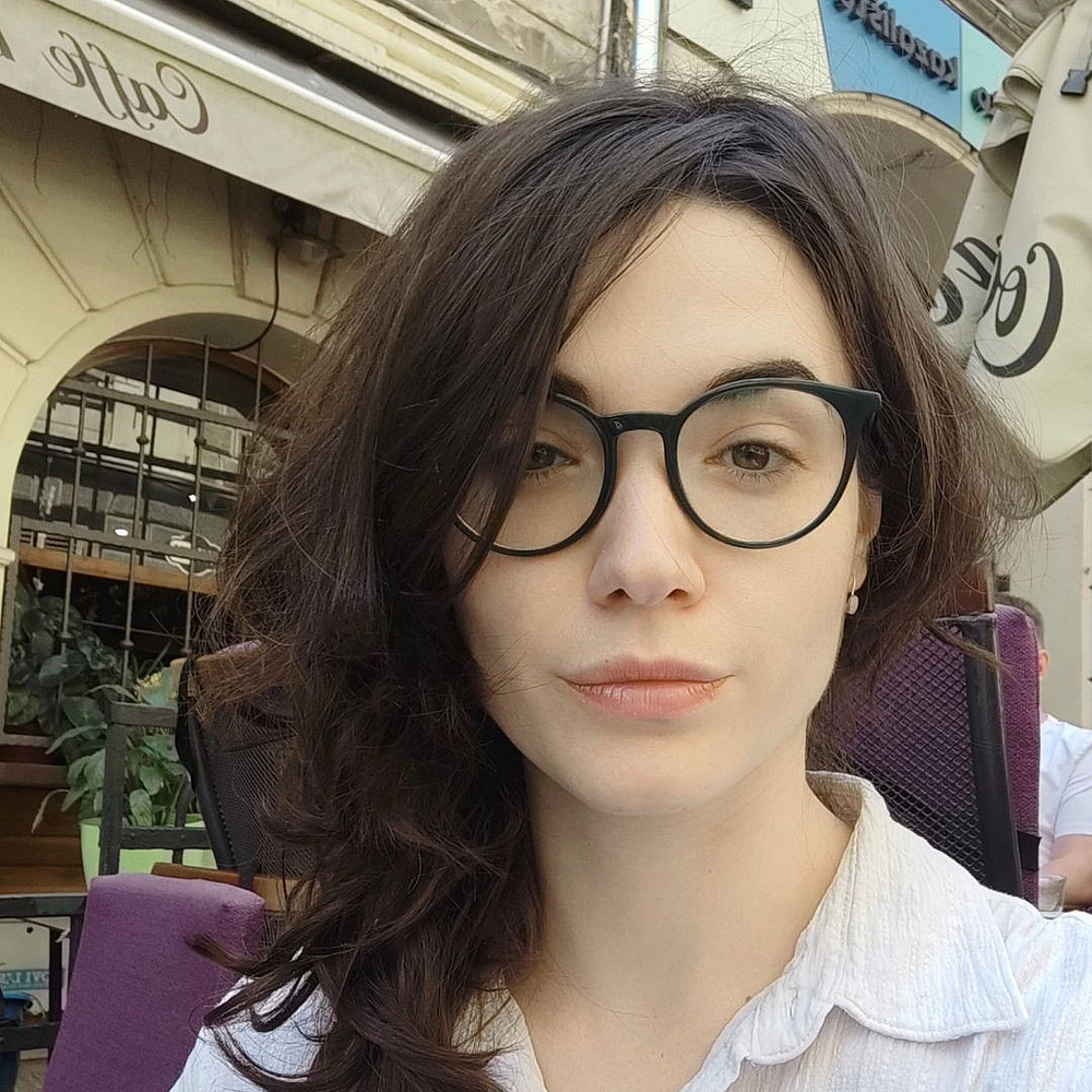 Das Foto zeigt Jelena Lalatović, eine Frau mit langen, dunklen Haaren und Brille, die direkt in die Kamera blickt. Sie trägt ein helles Hemd mit Knöpfen vor einem schlichten weißen Hintergrund, wodurch ein sanftes und minimalistisches Porträt entsteht. ©Jelena Lalatović