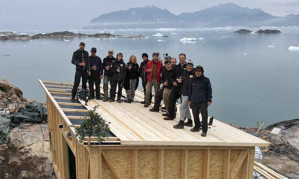 Ein Foto einer Gruppe von Menschen, die auf dem Dach eines kleinen Hauses stehen. Im Hintergrund sind das arktische Meer und Berge zu sehen, umgeben von zahlreichen Eisbergen. Die Menschen feiern ihre Leistung.