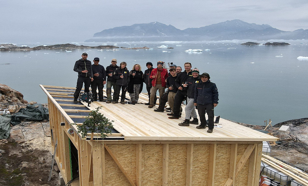 Ein Foto einer Gruppe von Menschen, die auf dem Dach eines kleinen Hauses stehen. Im Hintergrund sind das arktische Meer und Berge zu sehen, umgeben von zahlreichen Eisbergen. Die Menschen feiern ihre Leistung.