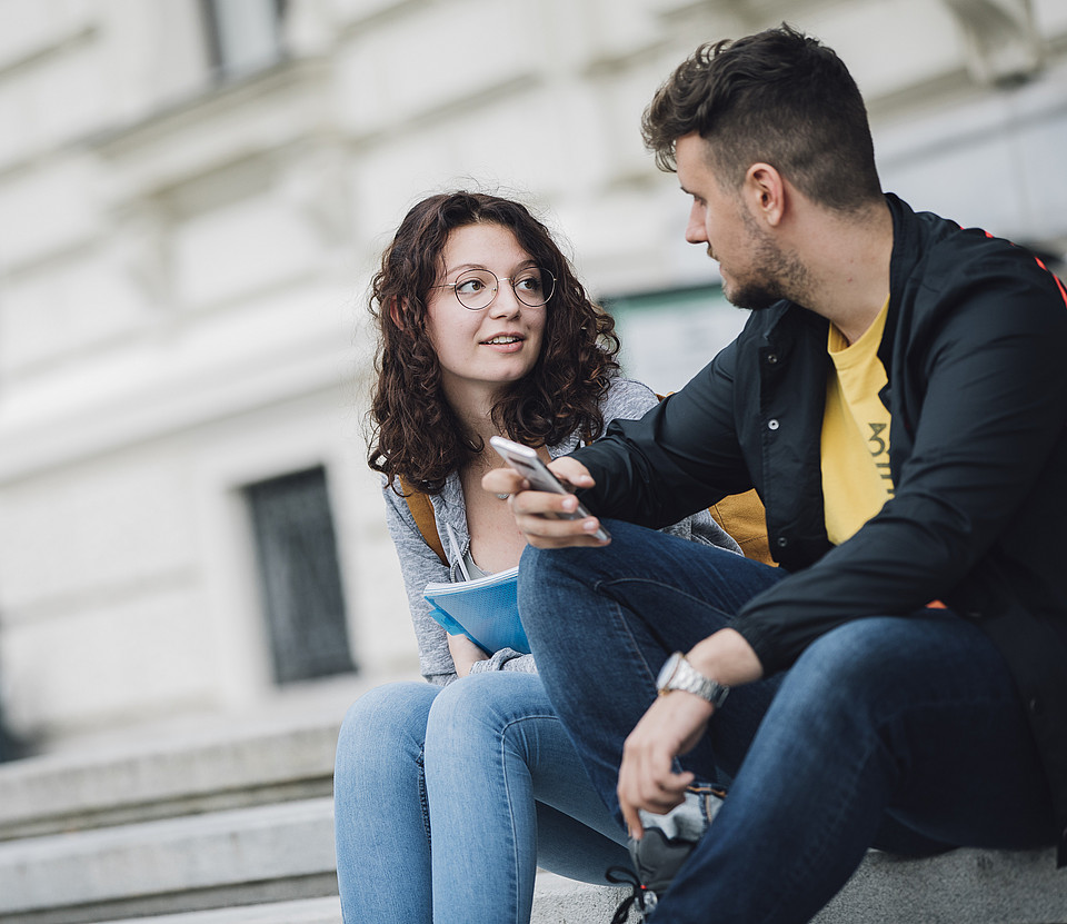 Zwei Studierende sitzen im Freien am Campusgelände