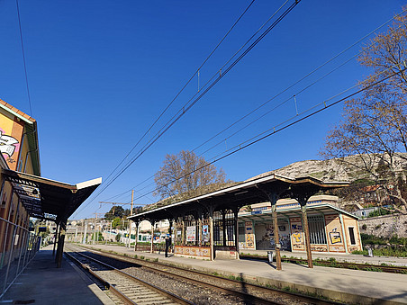 A quiet railway station with parallel train tracks, overhead power lines, and vintage metal platform shelters, set against a clear blue sky and a rocky hillside, with a few trees and graffiti-covered buildings nearby. ©Tijana Koprivica