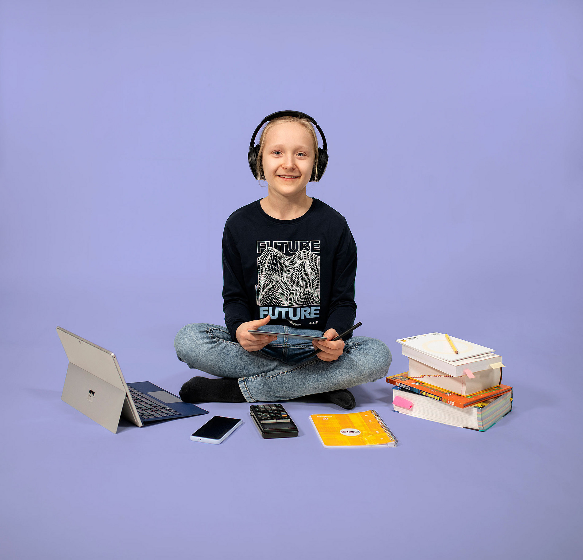 Ein Mädchen mit schwarzem, langärmlichen Shirt, Jeans und langen blonden Haaren sitzt mit Laptop, Handy, Taschenrechner, Block, und Büchern vor einem violetten Hintergrund. 