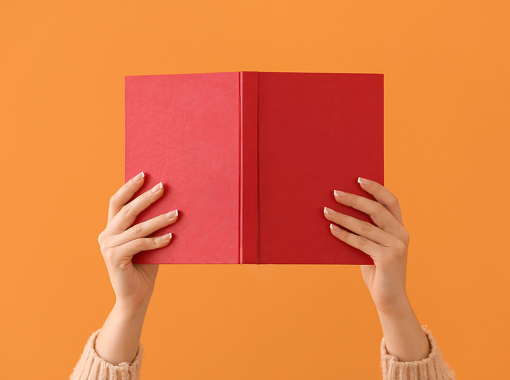 Information on submitting the exposé during the doctoral program 24W. A red book is held up by a woman in two hands against an orange background. ©pixel-shot.com (Leonid Yastremskiy)