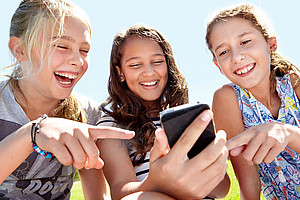 Drei lachende Mädchen, die auf ein Handy schauen. Das Mädchen in der Mitte hält das Handy in der Hand, die beiden anderen zeigen mit einem Finger darauf.