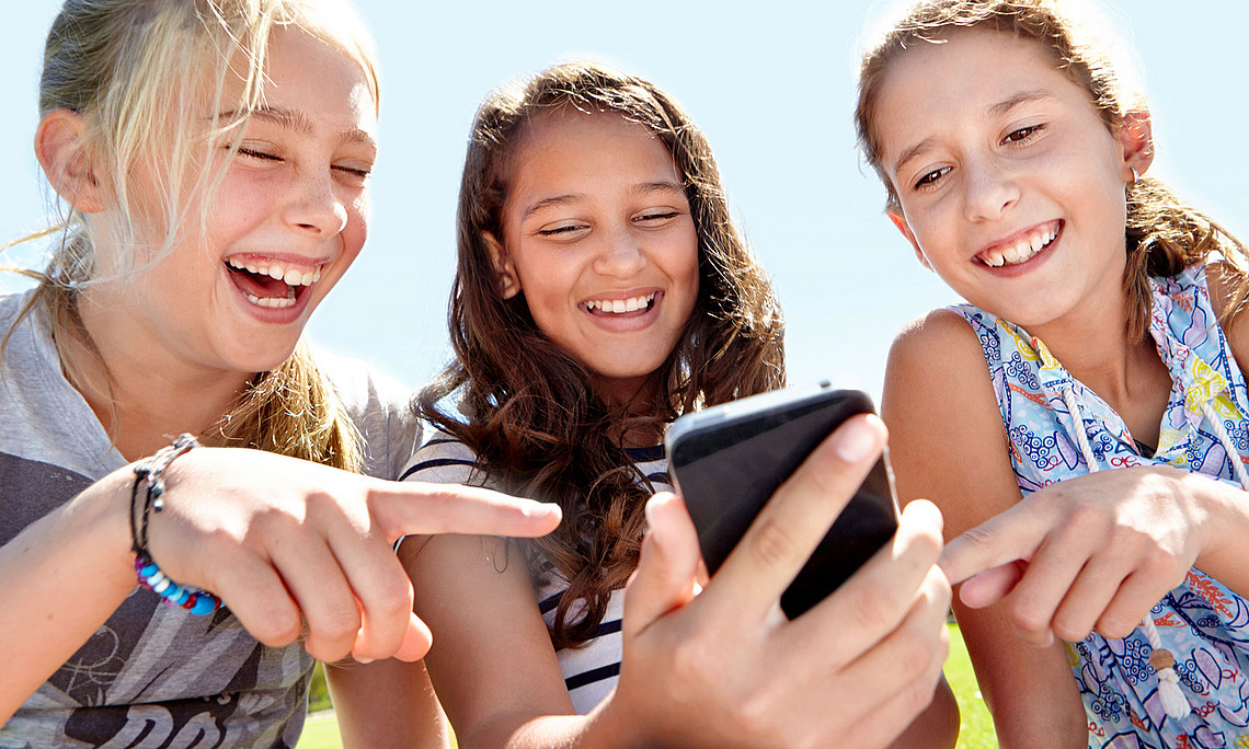 Drei lachende Mädchen, die auf ein Handy schauen. Das Mädchen in der Mitte hält das Handy in der Hand, die beiden anderen zeigen mit einem Finger darauf.
