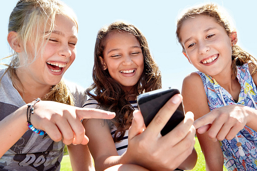 Drei lachende Mädchen, die auf ein Handy schauen. Das Mädchen in der Mitte hält das Handy in der Hand, die beiden anderen zeigen mit einem Finger darauf.