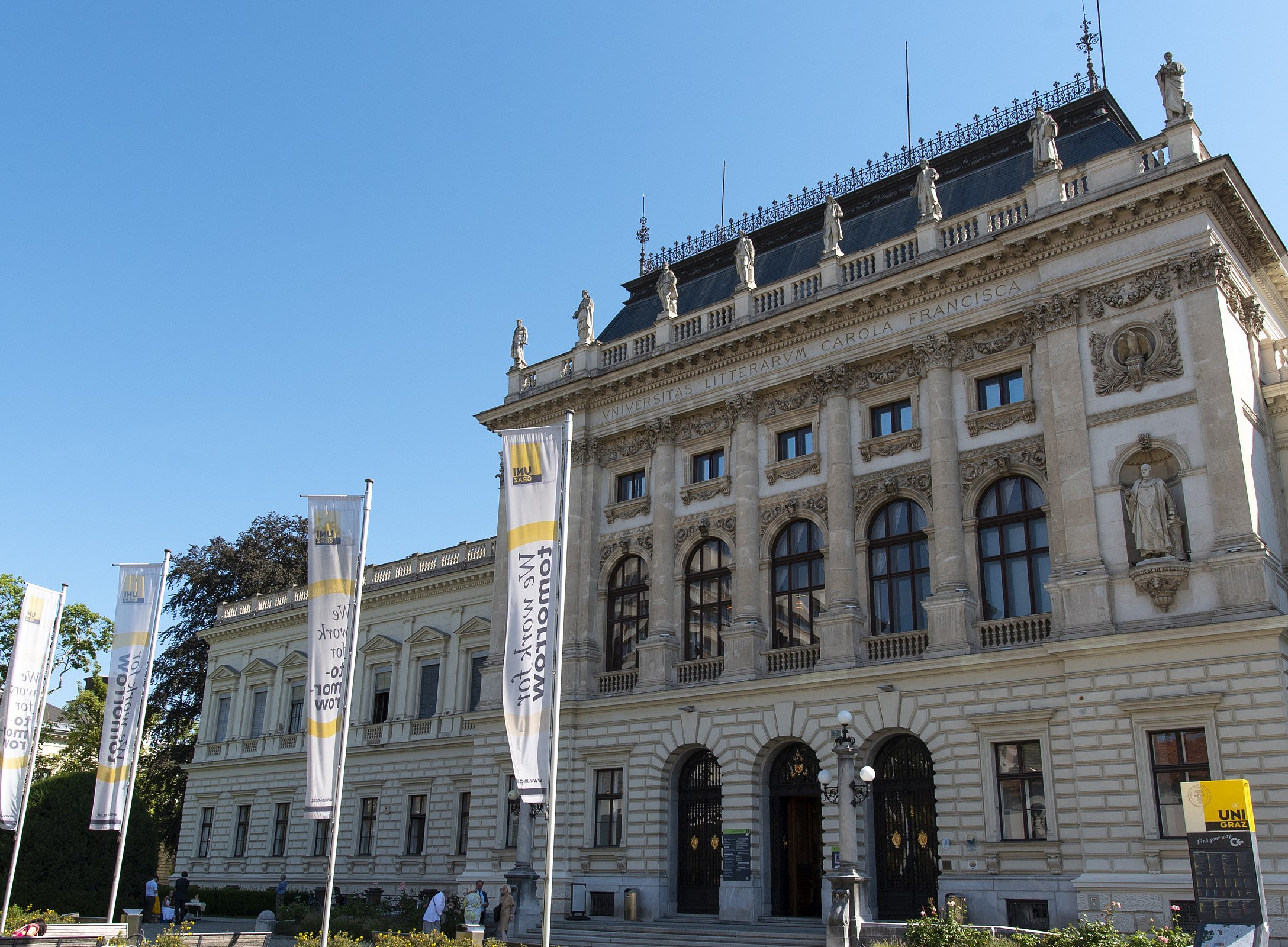 A magnificent, multi-storey historic building façade made of light-coloured stone, featuring tall arched windows, columns and statues in niches, with several tall flagpoles flying white and yellow flags in front of it. People are walking or sitting in the front garden area, which features flower beds and paths, against a clear blue sky. ©Universität Graz
