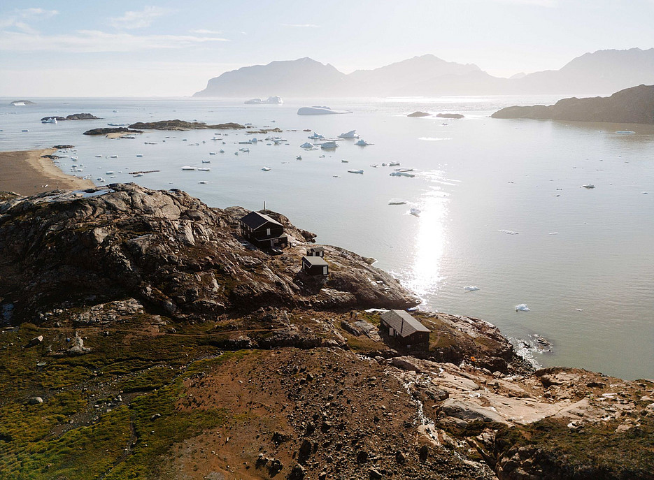 Blick von oben auf kleine Strukturen am Rande eines Strandes des Arktischen Ozeans, umgeben von großen schwimmenden Eisbergen und Inseln in der Ferne, mit einer üppig grünen Landschaft, die Sonne scheint, in diesem wachsenden Inselarchipel ©Karl Steinegger