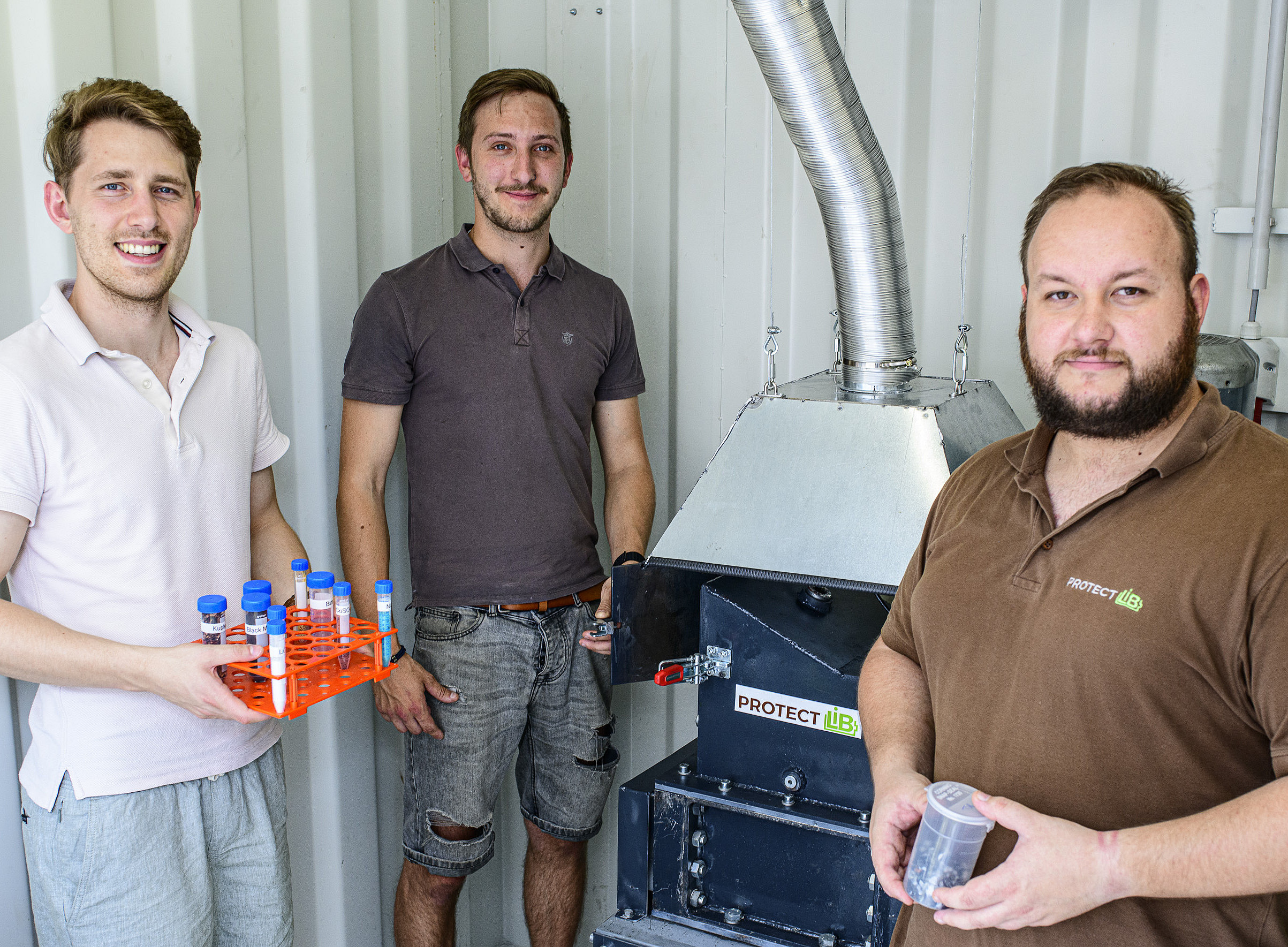 Drei Männer stehen neben einer Batterierecyclingmaschine im Hintergrund, die von hinten in einem Industriecontainer zu sehen ist Der Mann links hält Probenröhrchen mit verschiedenen Materialien in der Hand. Auf der einen Seite steht ein weiterer Mann in einem braunen T-Shirt, der einen durchsichtigen Becher mit geschreddertem Material hält. Der Mann in der Mitte trägt ein braunes T-Shirt und kurze Jeans. Sie posieren lächelnd für die Kamera, während sie in die Kamera schauen. ©Uni Graz/Tzivanopoulos