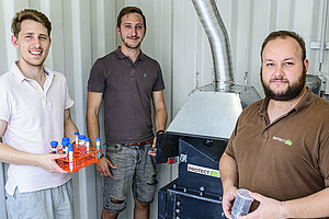 Drei Männer stehen neben einer Batterierecyclingmaschine im Hintergrund, die von hinten in einem Industriecontainer zu sehen ist Der Mann links hält Probenröhrchen mit verschiedenen Materialien in der Hand. Auf der einen Seite steht ein weiterer Mann in einem braunen T-Shirt, der einen durchsichtigen Becher mit geschreddertem Material hält. Der Mann in der Mitte trägt ein braunes T-Shirt und kurze Jeans. Sie posieren lächelnd für die Kamera, während sie in die Kamera schauen.