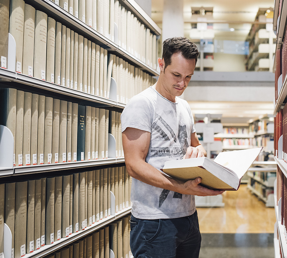 Ein Studierender blättert in einer Bibliothek zwischen zwei Bücherregalen in einem großen Buch