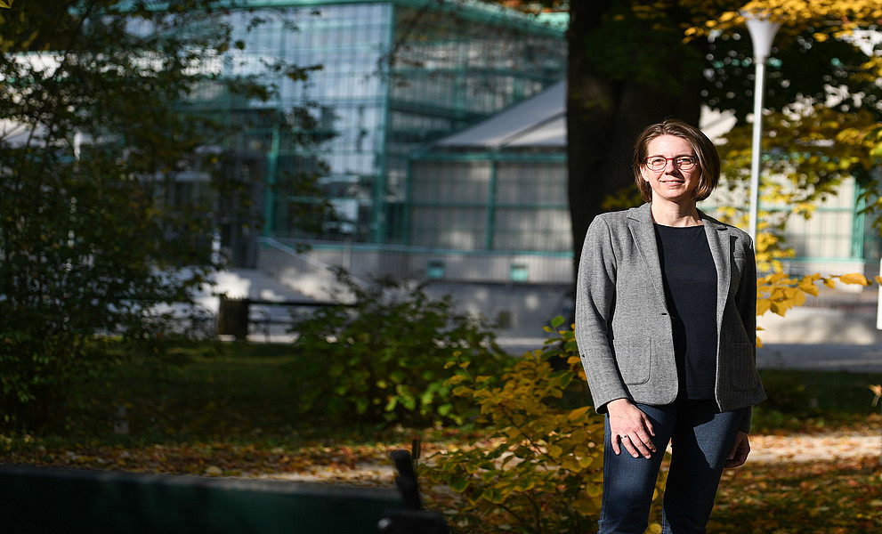 Erwachsene Frau mit kurzem Haar, Brille, grauem Blazer und Jeans steht im herbstlichen Park vor einem modernen Glasgebäude.