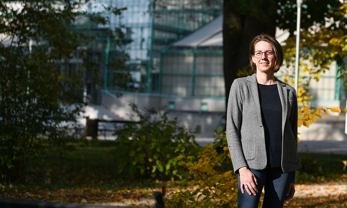 An adult woman with short hair, wearing glasses, a grey blazer and jeans, is standing in an autumnal park in front of a modern glass building.