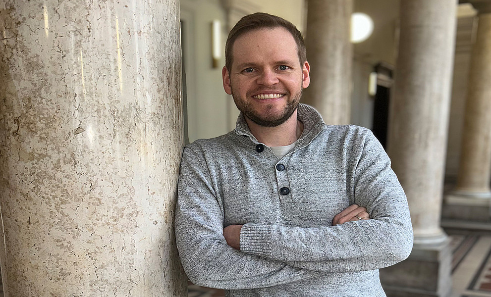 Dominik Sorger lehnt mit verschränkten Armen an einer Säule im Foyer des Uni-Graz-Hauptgebäudes