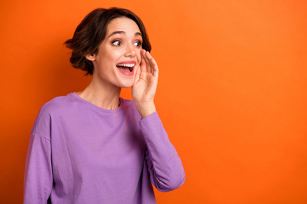 Woman in a purple sweater against an orange background holds her hand to her cheek and speaks ©deagreez - stock.adobe.com