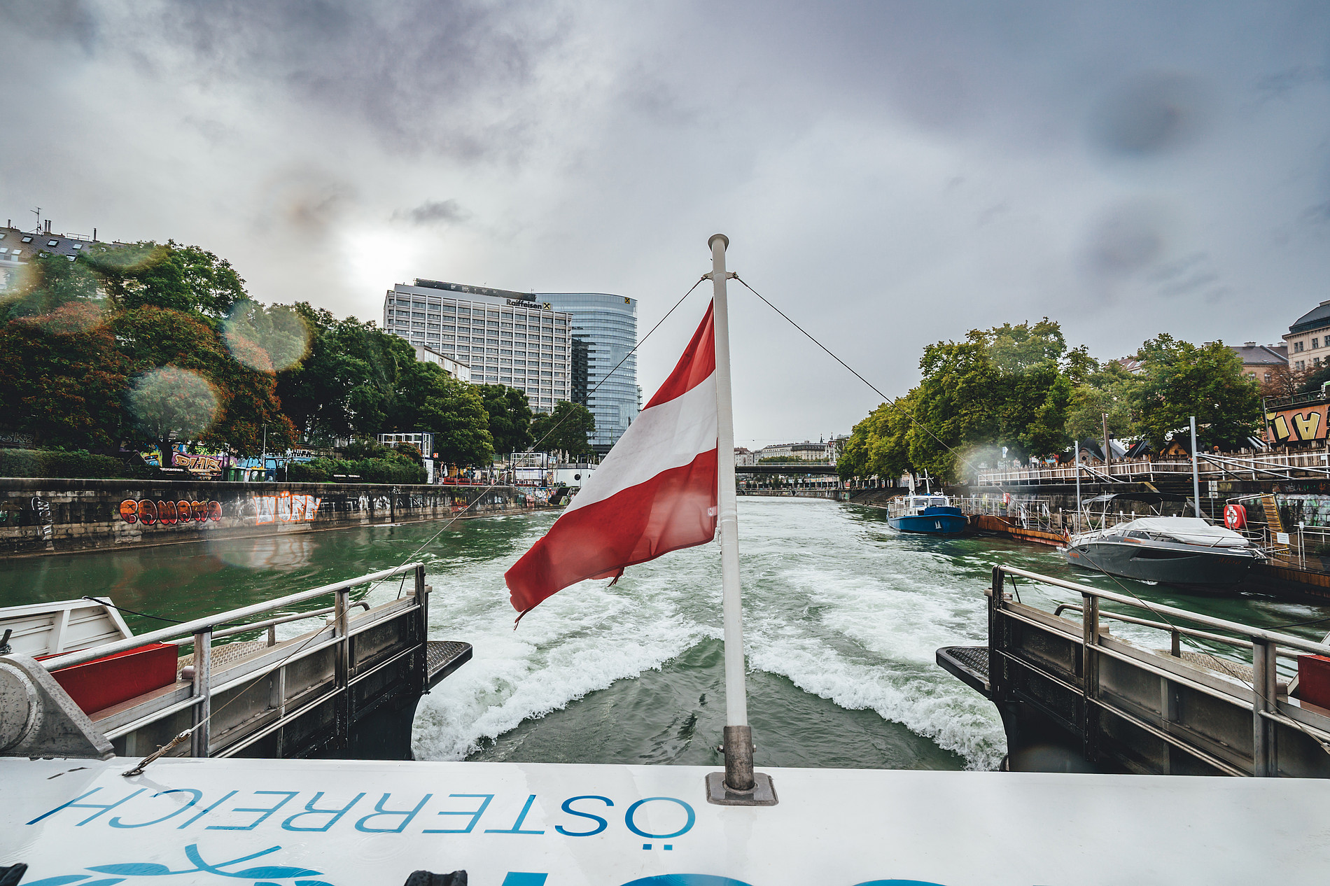 Austrian flag on a boat sailing down a city river, with modern buildings, trees, and other boats visible under a cloudy sky. ©Manuel Madaini
