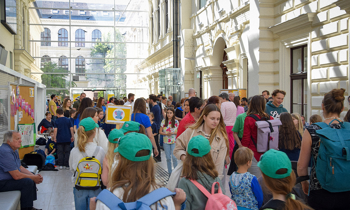 Gut besuchtes Foyer mit vielen Kindern, Lehrpersonen und Begleitpersonen, die zwischen Infoständen und bunten Plakaten einer Schul- oder Universitätsveranstaltung hindurchgehen.