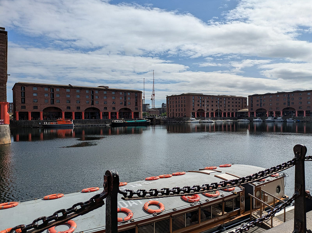 Das Foto zeigt einen weiten Blick auf das historische Albert Dock in Liverpool mit ruhigem Wasser, umgeben von großen Lagerhäusern aus rotem Backstein mit gewölbten Erdgeschossen. An den Rändern liegen Boote vor Anker, und im Vordergrund verläuft ein alter Kettenzaun. Der Himmel ist teilweise bewölkt, mit blauen Flecken und Sonnenlicht, das sich auf dem Wasser spiegelt. ©Daniella Shaw