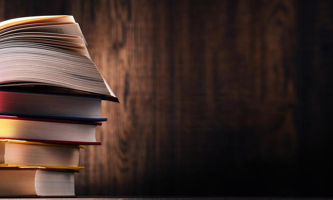 Ein Stapel Bücher, auf dem wiederum ein Buch liegt, welches aufgeschlagen ist.