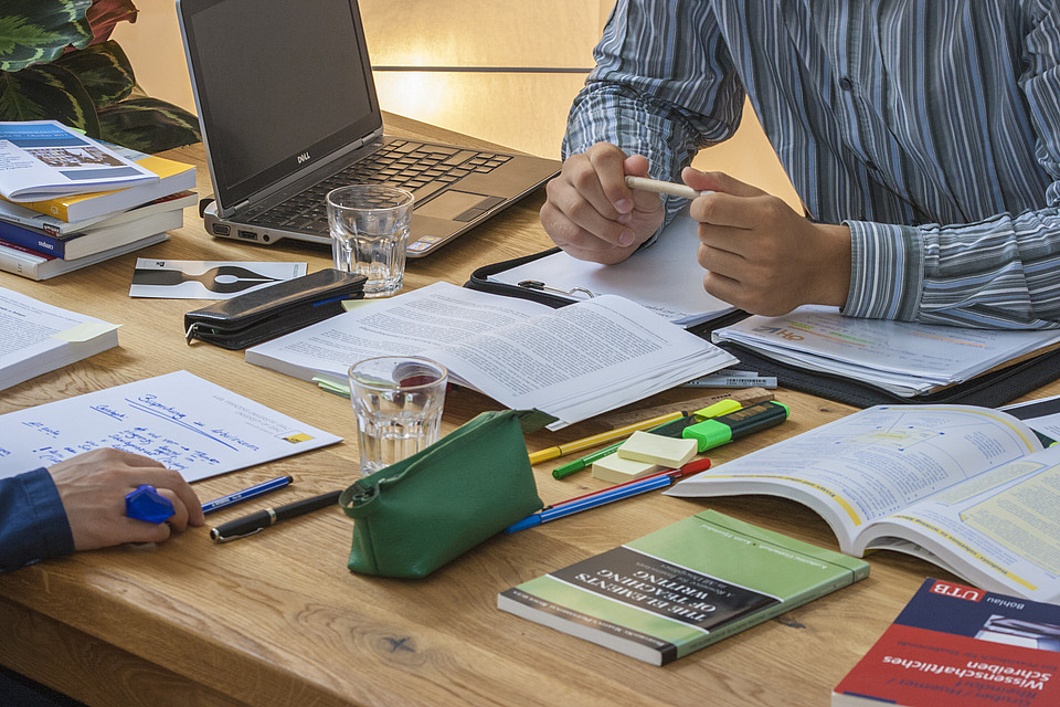 Tisch mit verschiedenen Schreibmaterialien, Wassergläsern, Stiften, Büchern, ein Flyer des Schreibzentrums. Zwei Personen sind teilweise zu sehen, die gerade miteinander über einen Text sprechen, der zwischen ihnen auf dem Tisch liegt. 